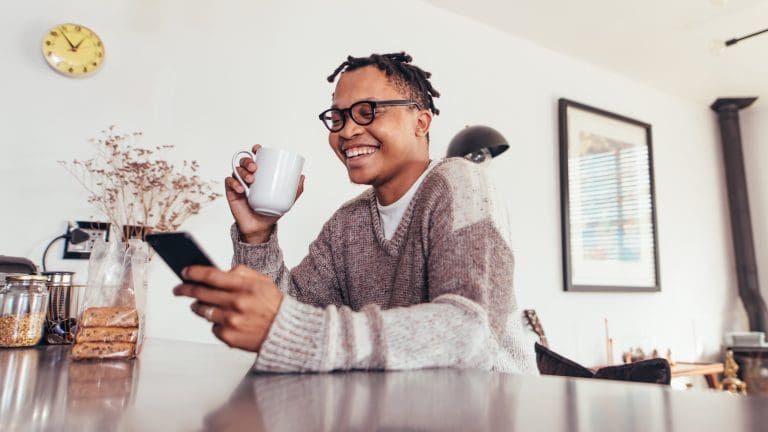 Happy young african man sitting at table using mobile phone and drinking coffee