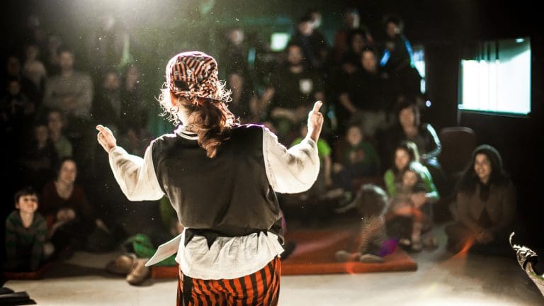 A performing arts entertainer is seen from the back in selective focus, dressed as a pirate on stage during a comedy act with blurry audience at back