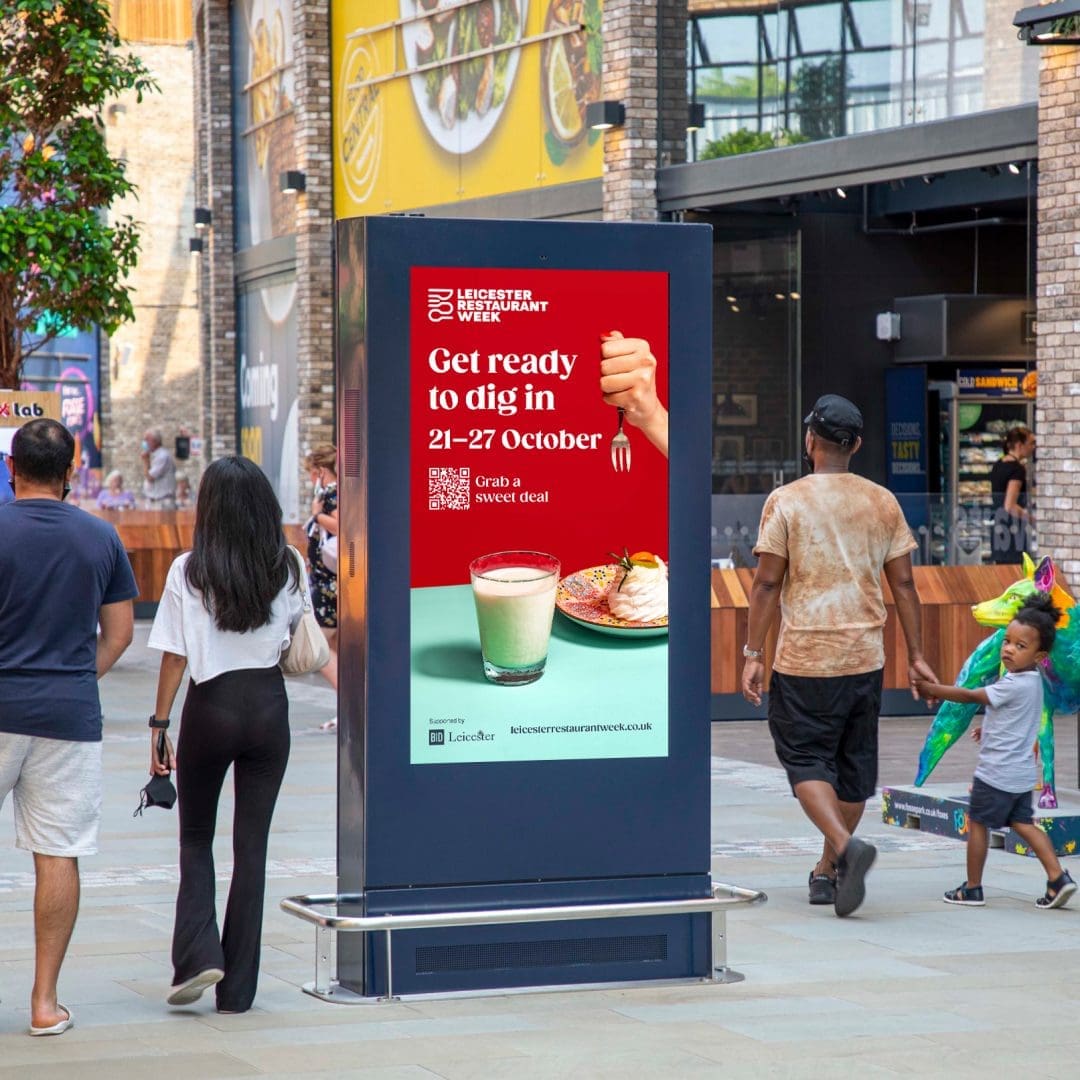 Family walking past a digital ad screen showing a Leicester Restaurant Week ad, with the tagline 'Get ready to dig in'.